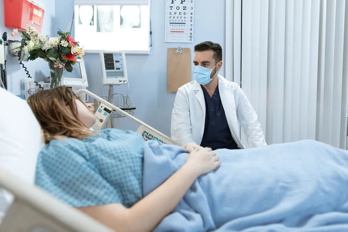 Médico de máscara conversa com uma paciente deitada em leito hospitalar durante a avaliação